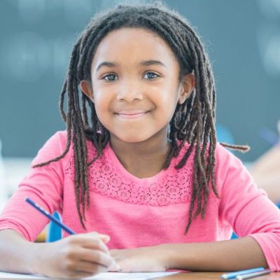 A young girl in a classroom with two other girls behind her. She has a colors pencil in her hand and she's smiling at the camera