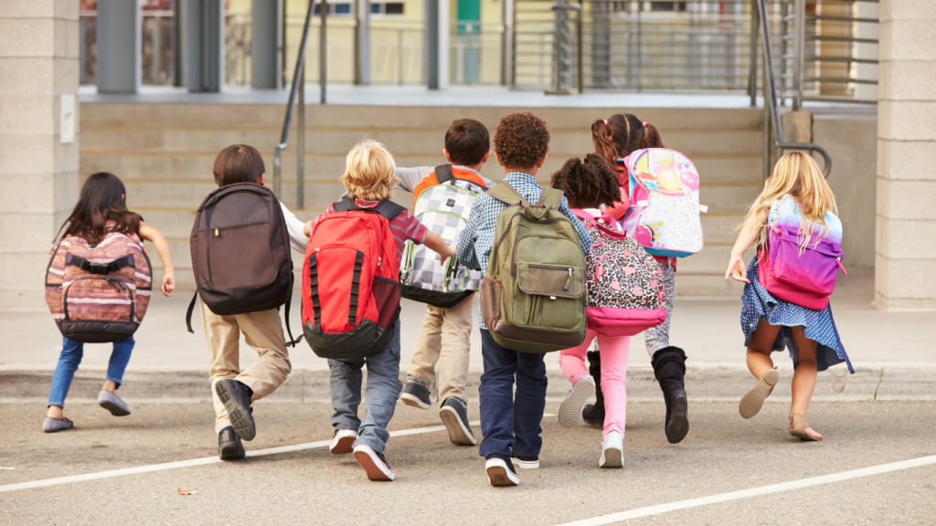 A group of school children with backpacks on. All of them are running towards a building.