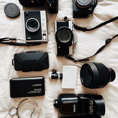 Camera gear laying on a flat surface. Sony camera body. Instax polaroid camera. Minolta film camera. Flashes and other gear.