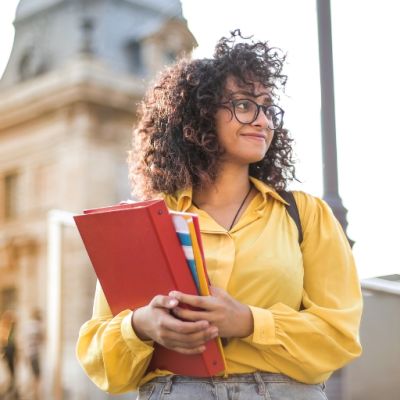Young college student with curly hair, wearing glasses and a yellow blouse. She is holding a stack of notebooks and folders. The background features a historic building, suggesting she is on a university campus. The student has a confident and focused expression.