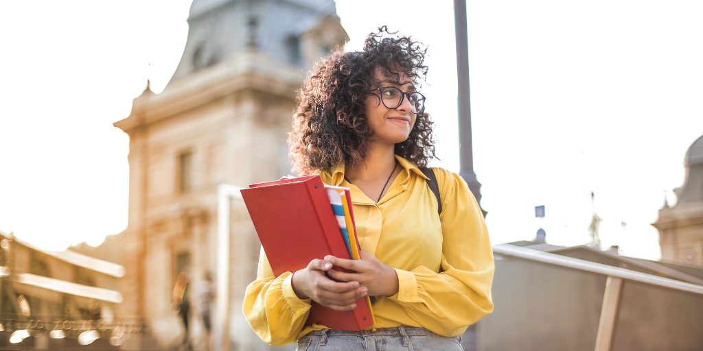 Young college student with curly hair, wearing glasses and a yellow blouse. She is holding a stack of notebooks and folders. The background features a historic building, suggesting she is on a university campus. The student has a confident and focused expression.