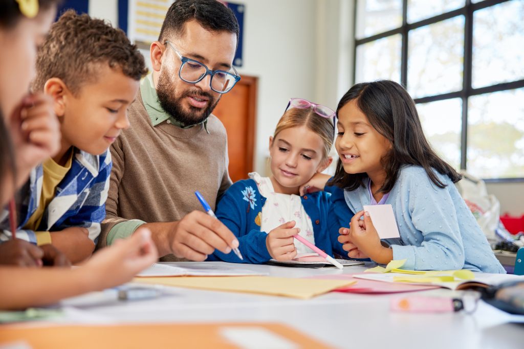 Teacher with glasses helps four children write on paper at classroom table.